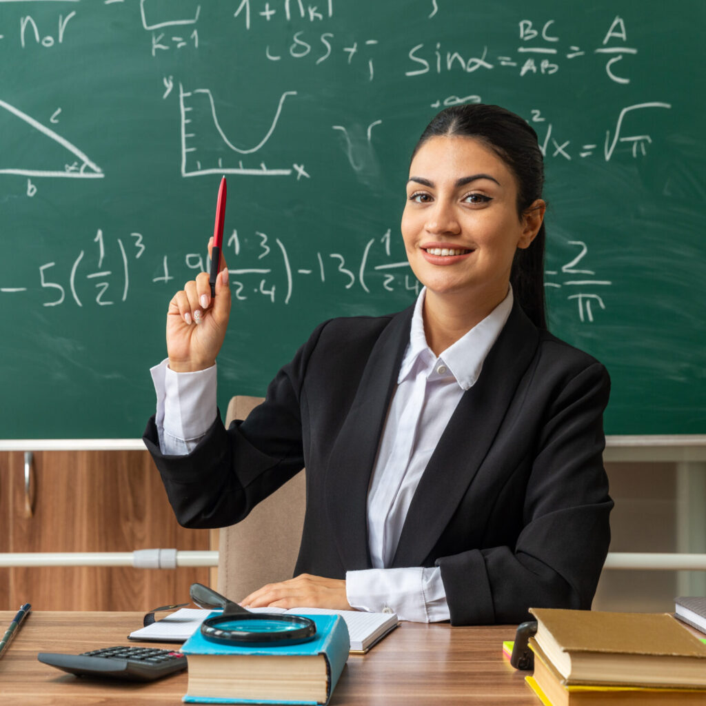 impressed young female teacher sits at table with school tools raising pen in classroom